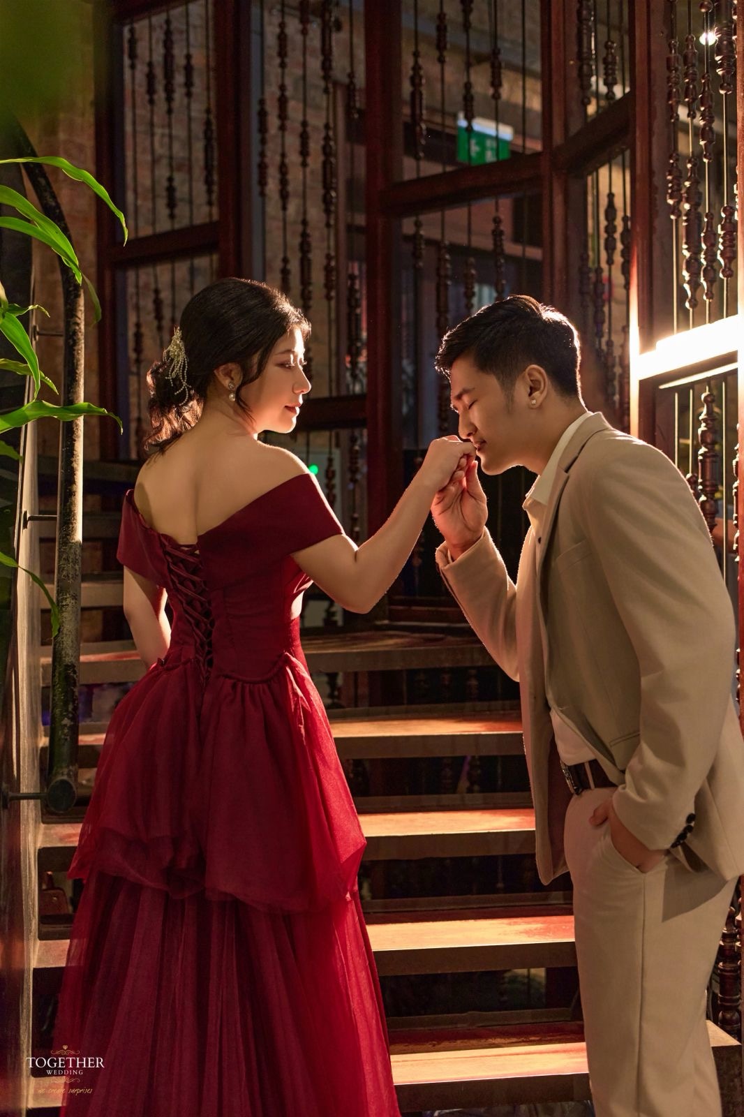 Mintzu and Zwe on a softly lit wooden staircase, Zwe kissing Mintzu's hand.